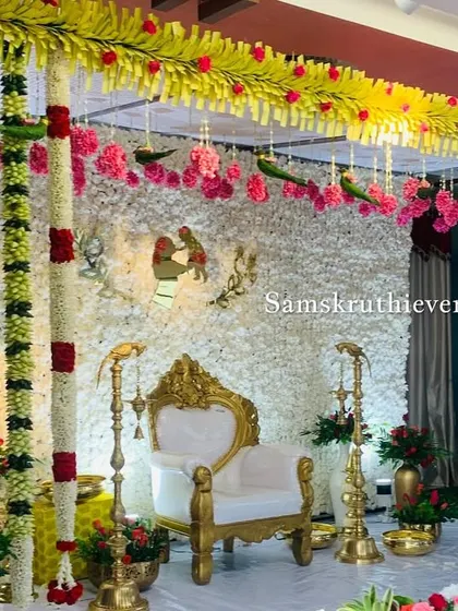 A traditional stage design with a white floral backdrop and hanging parrot motifs. The decor is framed by columns of jasmine and accented with red and green flowers.