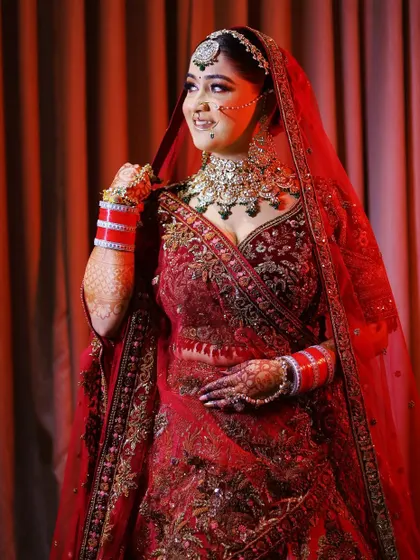 A stunning bridal portrait against a rich red background. The bride's confident pose and joyful expression perfectly match the vibrant color of her lehenga and the celebratory mood.