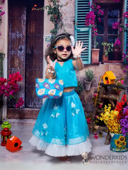 A little girl with a stylish attitude, wearing sunglasses and a modern blue lehenga in a colorful, European-inspired street scene. Her pose is full of confidence and fun.