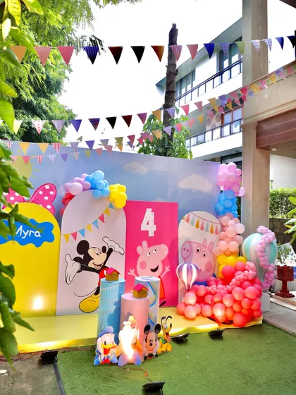 An outdoor party scene featuring colorful backdrops with Mickey Mouse and Peppa Pig. The festive bunting and balloon decorations complete this cheerful and fun-filled setup.