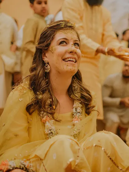 The bride's face, smeared with turmeric, is lit up with a beautiful, happy smile. This close-up captures the pure, unscripted joy of the Haldi ceremony.