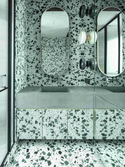 The younger daughter's bathroom is a playful mix of green and white terrazzo. A concrete wash basin, fluted terrazzo shutters, and hand-chiseled stone wall lights create a unique and vibrant space.