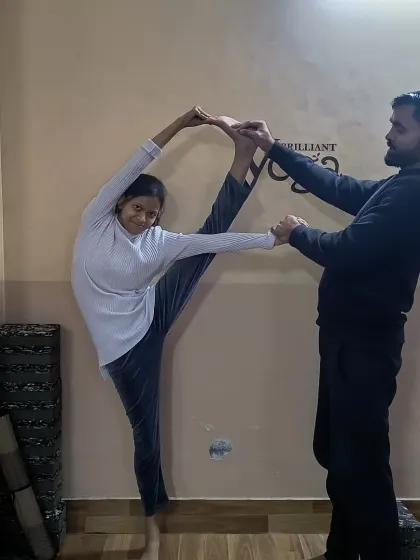 A student practices a variation of Trivikramasana, or Standing Splits, with instructor support. This pose deeply stretches the hamstrings and hips while challenging your balance and focus.
