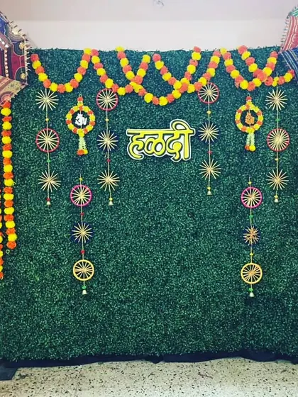 A beautiful Haldi decoration perfect for home celebrations. An artificial green wall is adorned with marigold garlands, a 'हळदी' sign, and colorful Rajasthani umbrellas, creating a perfect photo booth.