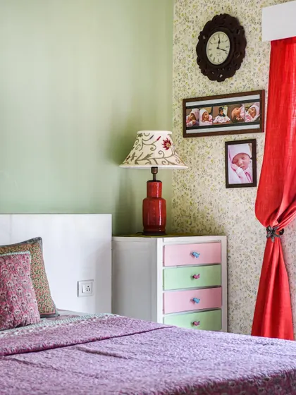 A child's room with soft green walls, a pop of color from the red curtains, and a chest of drawers with pastel-colored fronts.