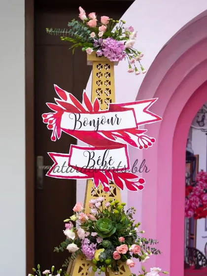A "Bonjour Bébé" signpost at a Paris-themed baby shower. The sign is part of a larger Eiffel Tower prop, adorned with flowers, creating a chic and thematic photo spot.