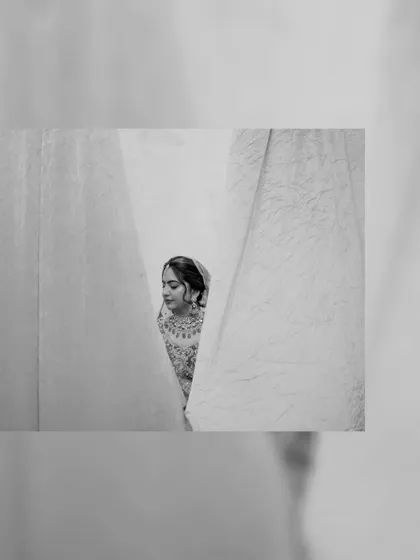 A candid black and white shot of the bride peeking through curtains, with a gentle smile.
