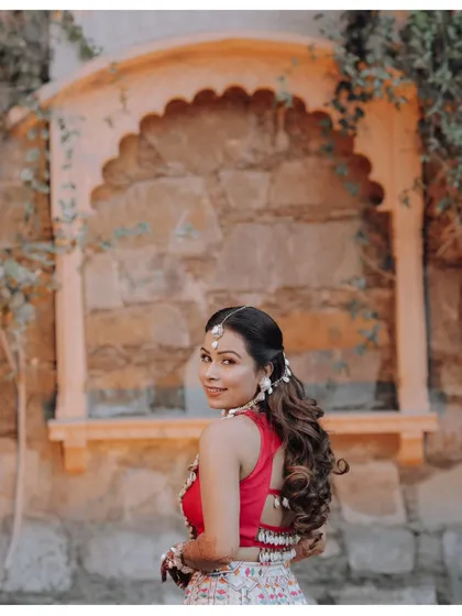 The bride looks over her shoulder with a smile, her intricate hairstyle and backless blouse featured against a rustic stone wall.