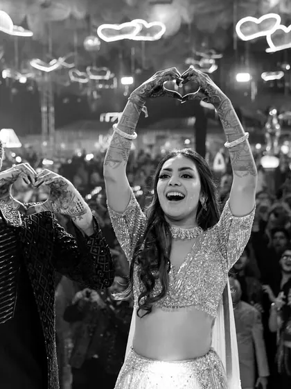 A heartwarming black and white shot of the couple making heart signs with their hands, sharing a moment of fun and love with their guests during the Sangeet.