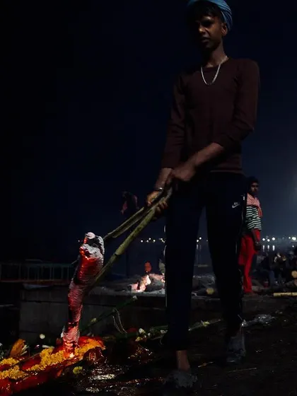 A young worker at Manikarnika Ghat uses long bamboo poles to tend to a funeral pyre at night. This candid action shot documents the difficult and essential work involved in the final rites.