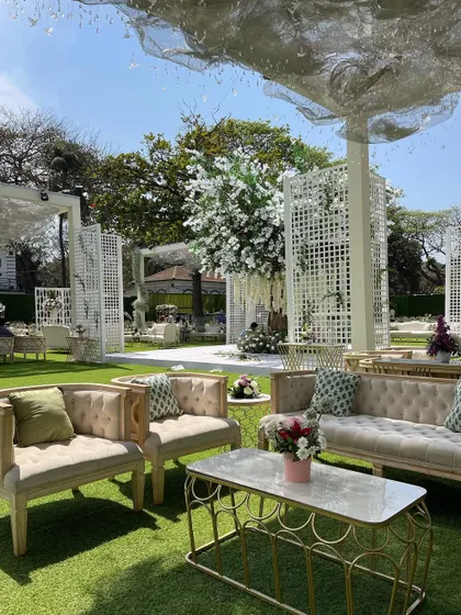 An elegant outdoor lounge area with a custom-built white floral tree as the centerpiece. The design combines comfortable seating with artistic floral installations for a relaxed yet sophisticated reception.
