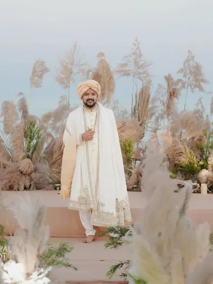The groom stands proudly at the pampas grass-adorned mandap during his sunset beach wedding ceremony.
