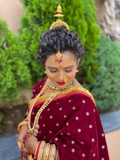 A traditional Maharashtrian bridal look. The hairstyle features tight curls forming a classic bun, adorned with traditional gold jewelry and a bindi for an authentic and elegant appearance.