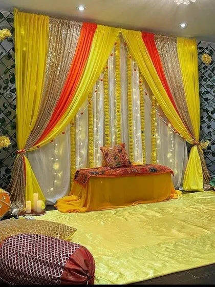 A beautiful Mehendi or Haldi seating area created with draped yellow, orange, and white fabrics. The backdrop is accented with sequin panels and lit with fairy lights, while floor cushions provide comfortable seating for guests.
