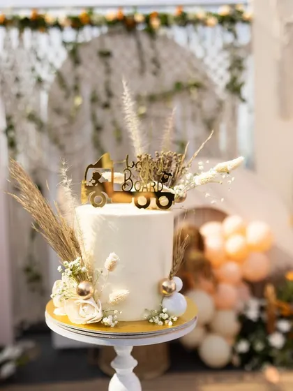 A beautiful cake for a boho-themed baby shower. The simple white cake is topped with dried grasses and a "Baby Boho" sign, perfectly matching the rustic and natural aesthetic.