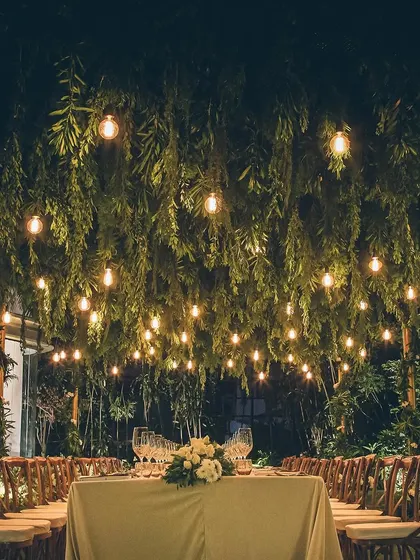 A magical outdoor dining setup with a long table under a canopy of hanging greenery and lights.