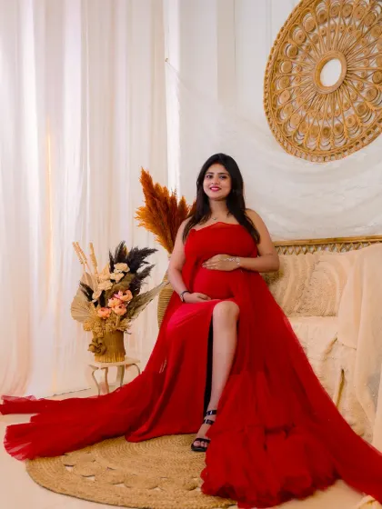 A portrait of pure elegance. The mom-to-be looks stunning in a bold red gown with a long, flowing train, seated in our bright and airy boho-themed studio setup.
