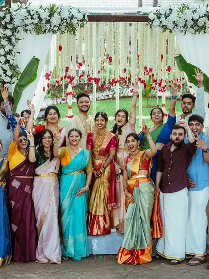 A cheerful group photo of the wedding party in their traditional attire, posing in front of an elegant floral mandap.