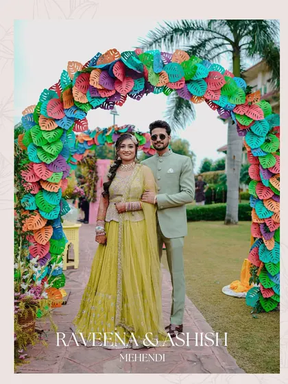 The couple poses under a vibrant, multi-colored leaf arch at their Mehendi ceremony. This shot highlights the unique and creative decor of the event.