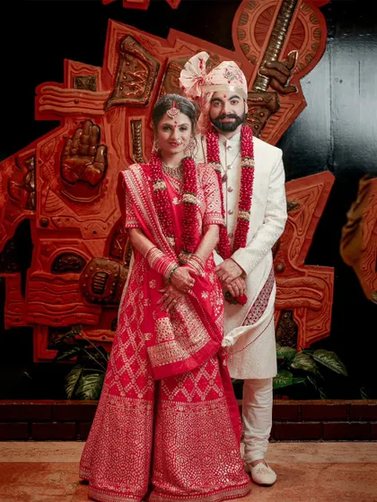A classic, full-length portrait of the bride and groom in their stunning red and white wedding attire, set against an artistic backdrop.