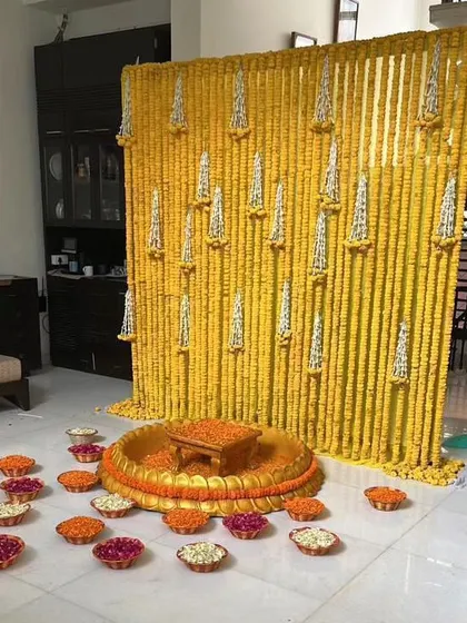 This indoor Haldi setup features a backdrop of yellow marigold strings with white floral accents. The large brass urli is filled with flower petals, ready for the rituals.