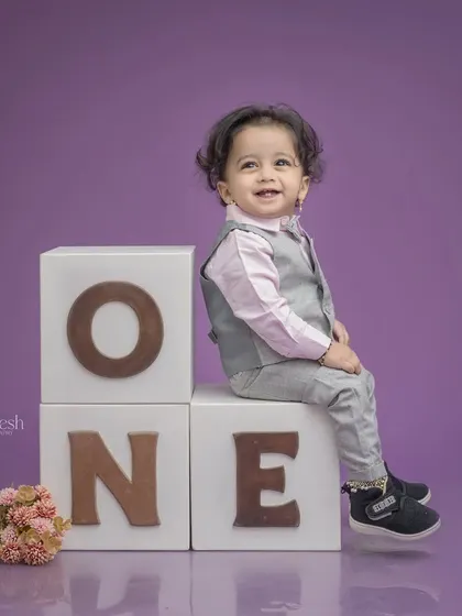 A dapper little gentleman celebrating his first birthday. He's sitting on 'ONE' blocks in a smart vest and shirt, giving a charming smile for his milestone portrait.