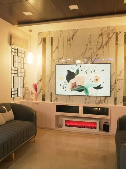 This luxurious living room in a Vaishnavi Commune apartment features a unique TV unit with an integrated electric fireplace. The marble-finish back panel and dark sofas with gold trim create a high-end, dramatic look.