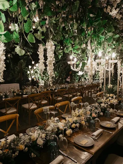 A close-up of the tablescape from a beautiful wedding dinner. The setting features elegant chandeliers dripping with white wisteria, creating a magical, intimate atmosphere for guests.