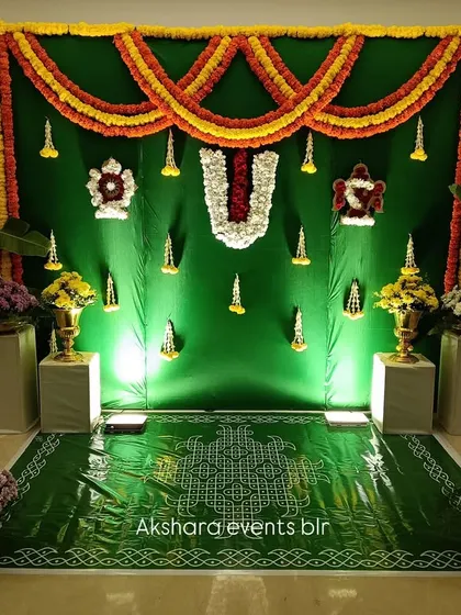 A simple yet elegant backdrop for a pooja ceremony. The green fabric is adorned with marigold garlands and a central 'Namam' symbol, with a traditional kolam drawn on the floor.