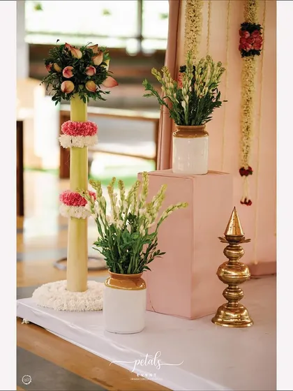 A corner of a ritual space, decorated with fragrant tuberoses in a pickle jar, a floral-wrapped banana stem, and a traditional brass Kalasa. This shows a creative mix of modern and traditional elements.