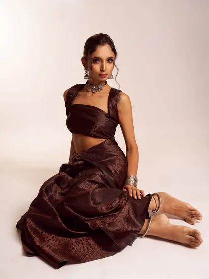 This full-length shot displays a unique, draped ethnic outfit. The makeup is kept strong yet simple, with defined eyes and a neutral lip to balance the rich brown fabric and statement silver jewelry.