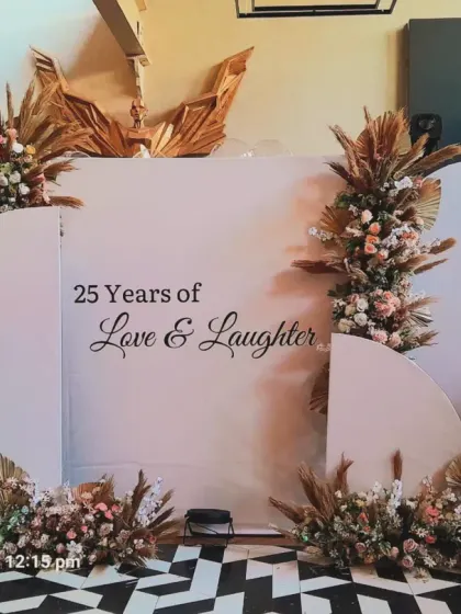 A modern and elegant backdrop celebrating "25 Years of Love & Laughter." The design uses clean arched panels and is decorated with boho-style pampas grass and floral arrangements.