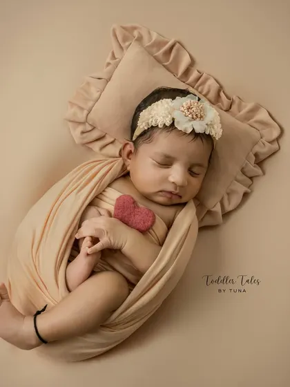 A 20-day-old baby girl, peacefully sleeping while wrapped in a soft peach swaddle. The tiny felt heart she holds adds a touch of sweetness to this classic newborn pose.