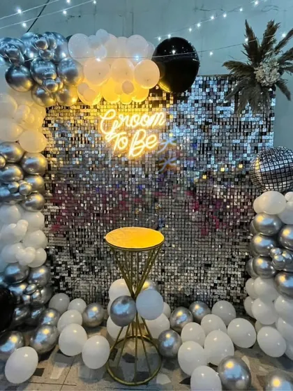 Celebrate the groom with a modern and stylish party setup. This "Groom to Be" decoration features a dazzling silver shimmer wall, a chic neon sign, and a sophisticated balloon arrangement in black, silver, and white, perfect for a bachelor party.