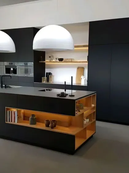 A minimalist kitchen design featuring matte black, floor-to-ceiling cabinetry and a matching island. The natural wood open shelving and warm lighting add contrast and prevent the space from feeling too stark.