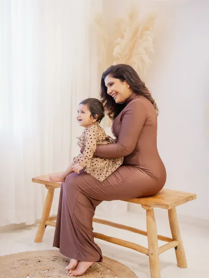 A sweet mother-daughter moment. The mom sits on a wooden bench, holding her one-year-old daughter on her lap, both sharing a happy and natural smile.