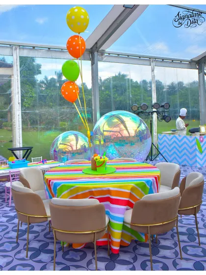 Guest seating at the summer carnival party, held inside a clear-span tent. The tables feature rainbow-striped linens and large, iridescent bubble balloons that catch the light beautifully.