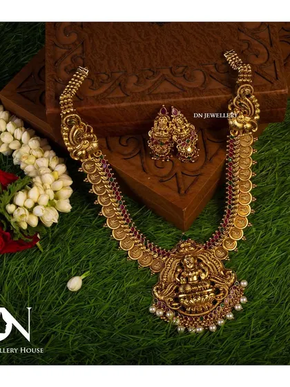 A beautiful temple necklace with a Lakshmi pendant, displayed on a bed of green grass. The natural setting highlights the antique gold finish.
