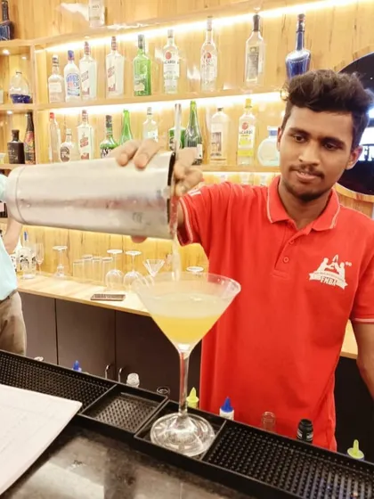 A student in a red polo shirt carefully pours a freshly shaken cocktail into a large martini glass during a practical class.