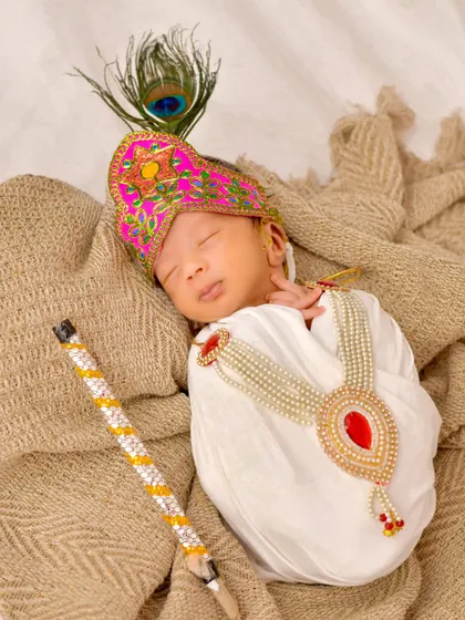 A precious newborn is dressed as little Krishna, complete with a peacock feather turban and flute, sleeping peacefully in a beautifully styled and traditional setup.