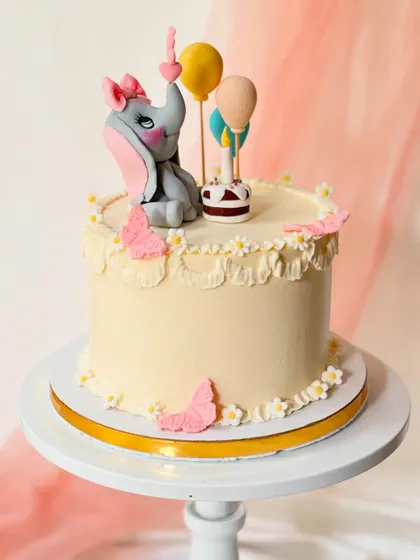 A sweet elephant-themed cake for a first birthday. The hand-sculpted elephant figure is holding a balloon, surrounded by butterflies and delicate buttercream piping.