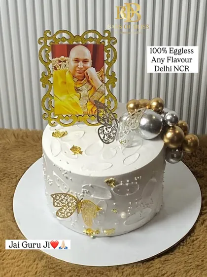 A cake made with devotion for a Guruji celebration. This elegant white cake is decorated with a photo of Guruji in a golden frame, delicate butterflies, and gold and silver spheres.
