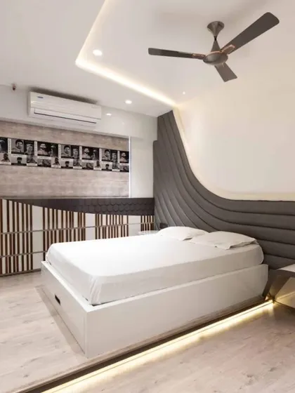 This bedroom is designed around the user's passion, with a custom headboard that flows into a bedside table. The room features a calm grey and white palette, bespoke furniture, and layered lighting for a sumptuous ambiance.