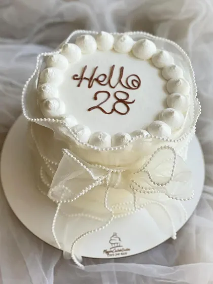 An elegant and simple "Hello 28" birthday cake. The all-white design is adorned with a delicate, translucent ribbon and bow.