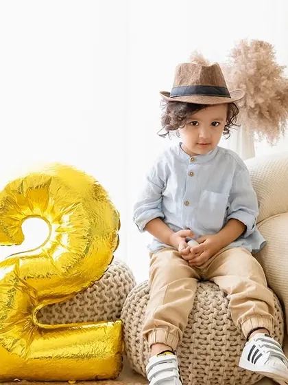 A toddler sits on a pouf next to a large golden "2" balloon for his second birthday photoshoot.