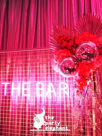 For this disco-themed event, I set up a stylish bar area with a neon 'THE BAR' sign against a hot pink backdrop. The look was completed with disco balls and red floral accents.