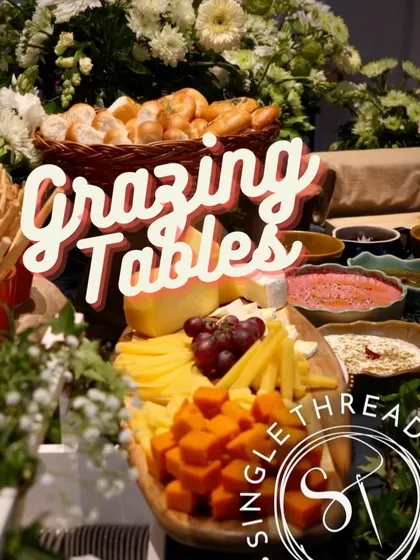 A branded look at one of my signature grazing tables. This image puts the focus on a beautiful wooden board laden with cubed and sliced cheeses, grapes, and a basket of bread, all framed by fresh flowers.
