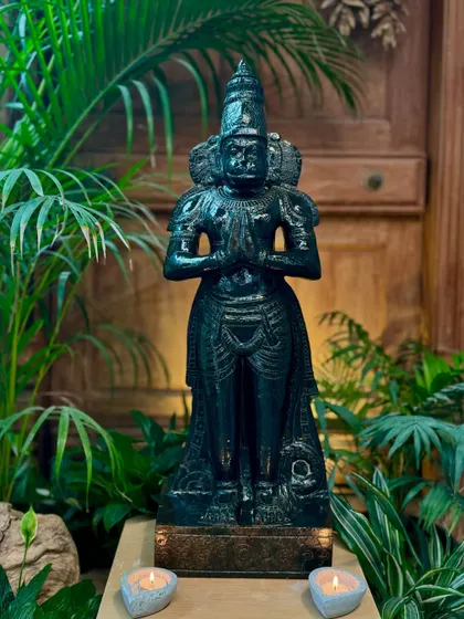 Our Anjali mudra Hanuman statue, carved from black granite, stands tall and serene in an indoor setting, flanked by plants and candles.