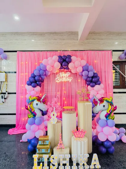 A clear, well-lit photo of the unicorn theme birthday decoration. The pink and purple balloon arch is perfectly formed around the pink curtain backdrop with fairy lights.