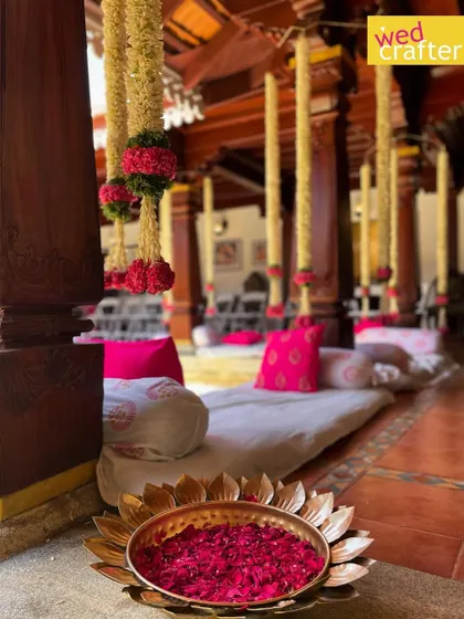 Details that honor tradition. A brass lotus bowl filled with fresh rose petals sits at the base of a wooden pillar, with jasmine and rose garlands hanging from the ceiling.